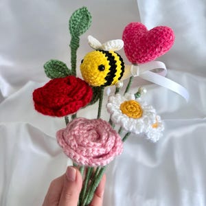 May include: A handmade crochet bouquet featuring a red rose, pink rose, white daisy, and a yellow and black bee. A pink heart and green leaves are also included. The bouquet is held in a hand.