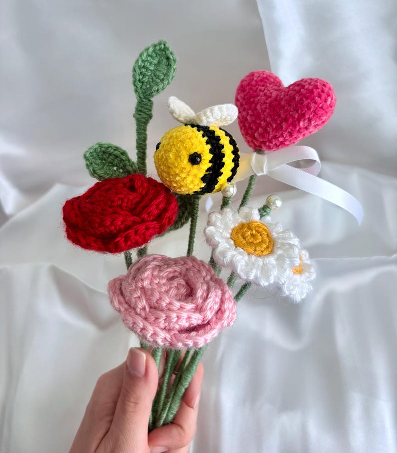 May include: A handmade crochet bouquet featuring a red rose, pink rose, white daisy, and a yellow and black bee. A pink heart and green leaves are also included. The bouquet is held in a hand.