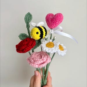 May include: A handmade crochet bouquet featuring a red rose, pink rose, white daisies, and a yellow and black bee. A pink heart with a white ribbon is also included. The bouquet is held in a hand.