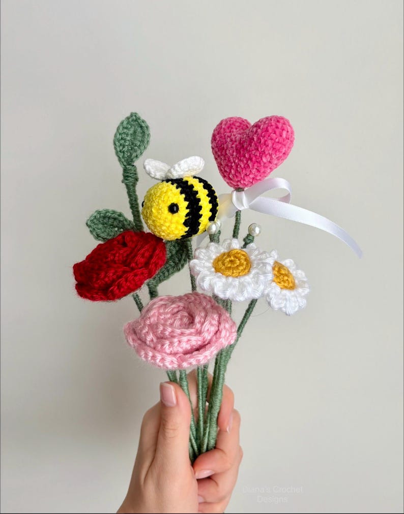 May include: A handmade crochet bouquet featuring a red rose, pink rose, white daisies, and a yellow and black bee. A pink heart with a white ribbon is also included. The bouquet is held in a hand.