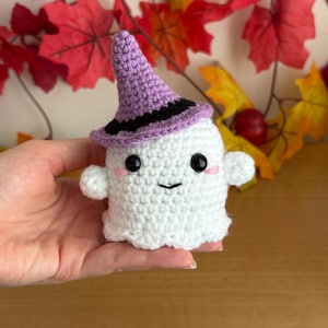 May include: A handmade white crochet ghost wearing a purple witch hat with a black band. The ghost has black eyes, a smiling mouth, and pink cheeks. The background features fall leaves.