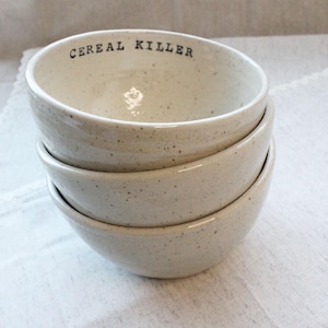 May include: A set of four white ceramic bowls with speckled glaze. The top bowl has the text "CEREAL KILLER" printed on the front.