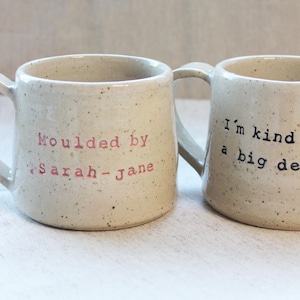 May include: Two white ceramic mugs with speckled glaze. One mug says "Moulded by Sarah-Jane" in pink lettering. The other mug says "I'm kind of a big deal" in black lettering.