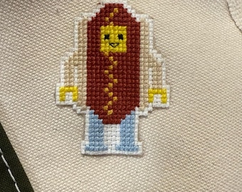 May include: A pixelated hot dog patch with a red body, yellow face, and blue pants. The hot dog has a yellow mustard design. The patch is on a cream-colored canvas bag.