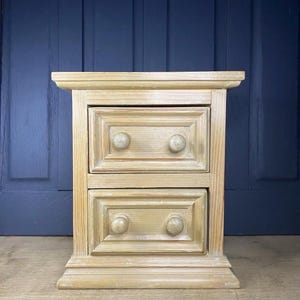 May include: A two-drawer bedside cabinet made of light-coloured wood. Each drawer features two round knobs. The cabinet has a rectangular form with a slightly projecting top and a base that widens at the bottom. The wood grain is visible.