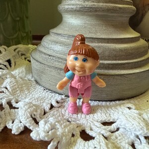 May include: A small plastic doll with brown hair styled in a high ponytail and bangs. The doll has blue eyes, a smiling expression, and wears a light blue short-sleeved shirt under pink overalls with matching pink shoes on its feet.