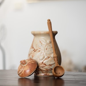 Könnte beinhalten: Eine beige Keramikvase mit Deckel und einem Holzlöffel. Die Vase hat ein Blumendesign und befindet sich neben ihrem Deckel. Der Holzlöffel lehnt an der Vase. Die Gegenstände befinden sich auf einer dunkelbraunen Oberfläche.