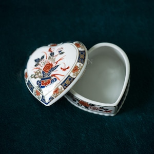 May include: Heart-shaped trinket box with a floral design. The lid is open, revealing the interior. The box is white with a detailed pattern of red, blue, and gold accents. The box is on a dark green surface.