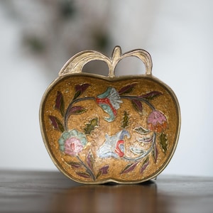 Könnte beinhalten: Goldfarbene, apfelförmige Zierschale mit Stielgriff. Die Schale zeigt ein Blumenmuster in Blau, Rosa und Grün auf goldenem Grund. Wahrscheinlich aus Metall gefertigt, eignet sie sich für dekorative Zwecke und hat ein Vintage-Design.