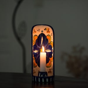 May include: A ceramic candle holder with a floral design in blue, orange, and yellow. A lit white candle sits inside the holder, casting a warm glow. The holder is on a dark wooden surface.