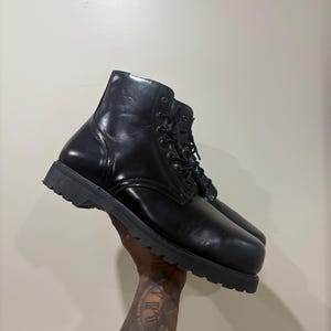 Könnte beinhalten: Ein schwarzer Lederstiefel mit glatter, polierter Oberfläche, abgerundeter Zehenpartie, Schnürung mit schwarzen Ösen und passenden schwarzen Schnürsenkeln sowie einer dicken, profilierten schwarzen Gummisohle für einen festen Halt.