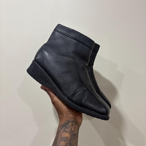 May include: A pair of black ankle boots featuring a smooth leather-like finish, a rounded toe, and a wedge heel. The boots have a clean, minimalist design with a visible seam running down the centre of the front and a structured collar at the top.