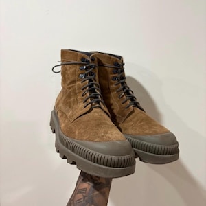 May include: A pair of brown suede ankle boots featuring dark brown laces, metal eyelets, and a thick, ribbed olive-green rubber lug sole that extends around the toe cap. The boots have a high-top silhouette with a textured, matte finish.