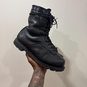 Puede incluir: Una bota alta de cuero negro con punta redondeada, suela de goma gruesa con tacos y cierre de cordones con ojales metálicos. La bota presenta un acabado liso, un cuello acolchado y cordones negros atados en la parte superior del tobillo.