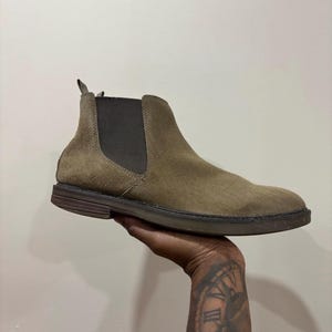 May include: A tan suede Chelsea boot featuring a dark brown elastic side panel, a pull tab at the heel, and a dark brown rubber sole. The boot has a rounded toe and a low heel, showcasing a textured suede finish across the upper portion of the shoe.
