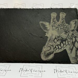 May include: A black slate serving tray featuring a laser-etched design of a giraffe's head. The giraffe is facing the right side of the tray and has a detailed pattern of spots.