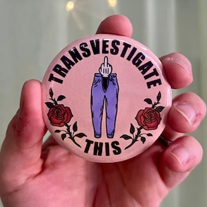 May include: Pink button with the text "Transvestigate This" in black lettering. A middle finger is sticking out of the top of a pair of blue jeans. Two red roses are on either side of the jeans.