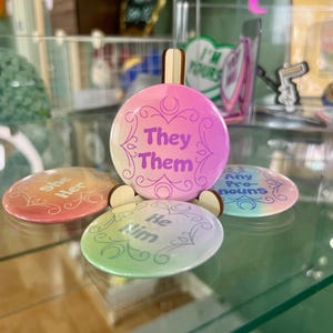 May include: Four round buttons with different pronoun options. The buttons are pink, orange, green, and blue with white text. The text on the buttons reads "She Her", "They Them", "He Him", and "Any Pronouns".