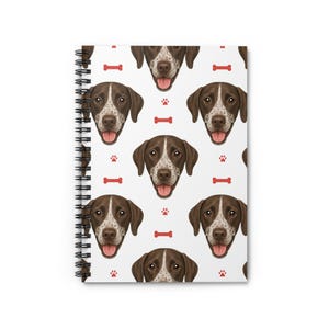May include: A spiral-bound notebook with a repeating pattern of a dog's face, red paw prints, and red dog bones on a white background. The dog has brown and white fur with its tongue out. The notebook's spiral binding is black.