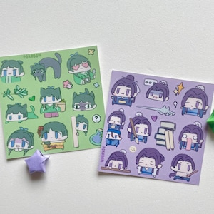 May include: Two sticker sheets with cartoon illustrations. One sheet is green with various characters and a black cat. The other sheet is purple with different character poses. A green and a purple origami star are also present.