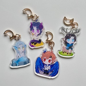 Puede incluir: Cuatro llaveros de personajes coloridos con anillos de metal dorado. Cada llavero presenta un personaje diferente con características y colores únicos. Los personajes incluyen una sirena, una figura alada, un demonio y una persona-ciervo.