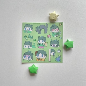 May include: A sticker sheet with various cartoon character designs in green, blue, and yellow. The sheet includes images of a cat, hearts, and other decorative elements. The word "POKOBON" is printed at the top. Three origami stars in yellow and green are scattered around the sticker sheet.