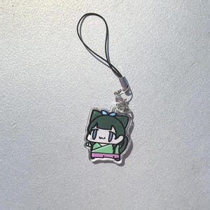 May include: A clear acrylic charm featuring a cartoon character with green hair and a blue bow. The character wears a green and pink outfit. The charm is attached to a black cord with a silver-colored clasp.