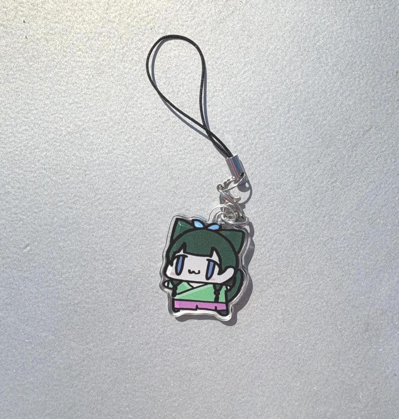 May include: A clear acrylic charm featuring a cartoon character with green hair and a blue bow. The character wears a green and pink outfit. The charm is attached to a black cord with a silver-colored clasp.