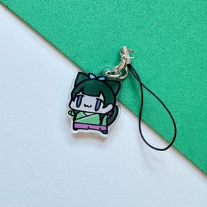 May include: A clear acrylic charm featuring a cartoon character with green hair and a blue bow. The charm has a silver-colored clasp and a black lanyard. The character is wearing a green and pink outfit. The background is white and green.