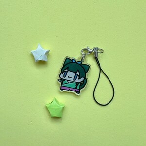 May include: A clear acrylic charm featuring a cartoon character with green hair and a blue bow. The charm is attached to a silver clasp and black string. Two origami stars, one white and one green, are also present on a yellow background.