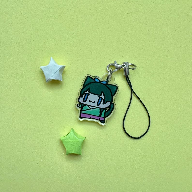 May include: A clear acrylic charm featuring a cartoon character with green hair and a blue bow. The charm is attached to a silver clasp and black string. Two origami stars, one white and one green, are also present on a yellow background.
