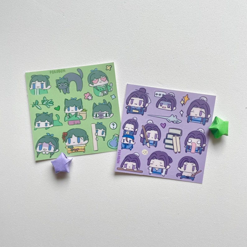 May include: Two sticker sheets with cartoon illustrations. One sheet is green with various characters and a black cat. The other sheet is purple with different character poses. A green and a purple origami star are also present.