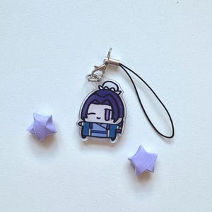 May include: A clear acrylic charm featuring a cartoon character with purple hair and blue clothing. The charm has a silver clasp and a black lanyard. Two small, folded purple paper stars are also present.