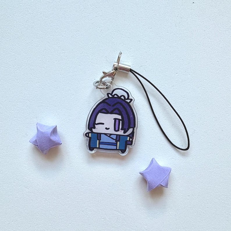 May include: A clear acrylic charm featuring a cartoon character with purple hair and blue clothing. The charm has a silver clasp and a black lanyard. Two small, folded purple paper stars are also present.