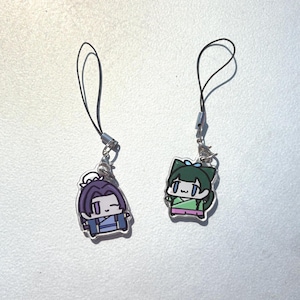 May include: Two cartoon character charms with black lanyards. One charm features a purple-haired character in blue clothing. The other charm depicts a green-haired character in green and pink clothing. Both charms have silver-colored metal clasps.