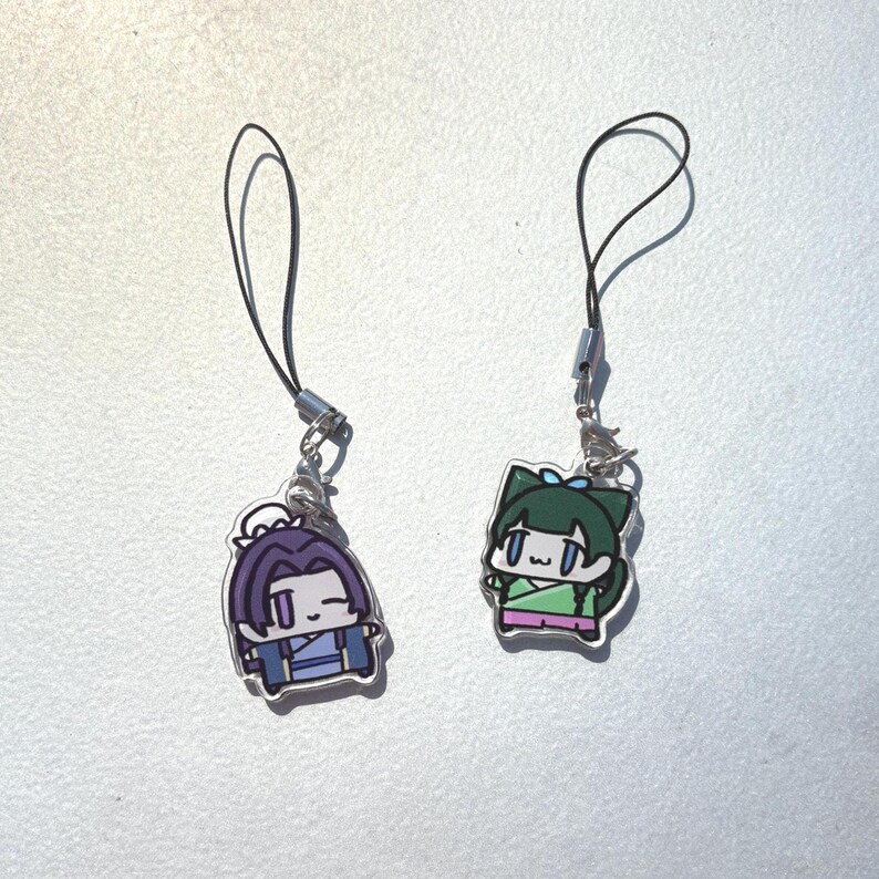 May include: Two cartoon character charms with black lanyards. One charm features a purple-haired character in blue clothing. The other charm depicts a green-haired character in green and pink clothing. Both charms have silver-colored metal clasps.