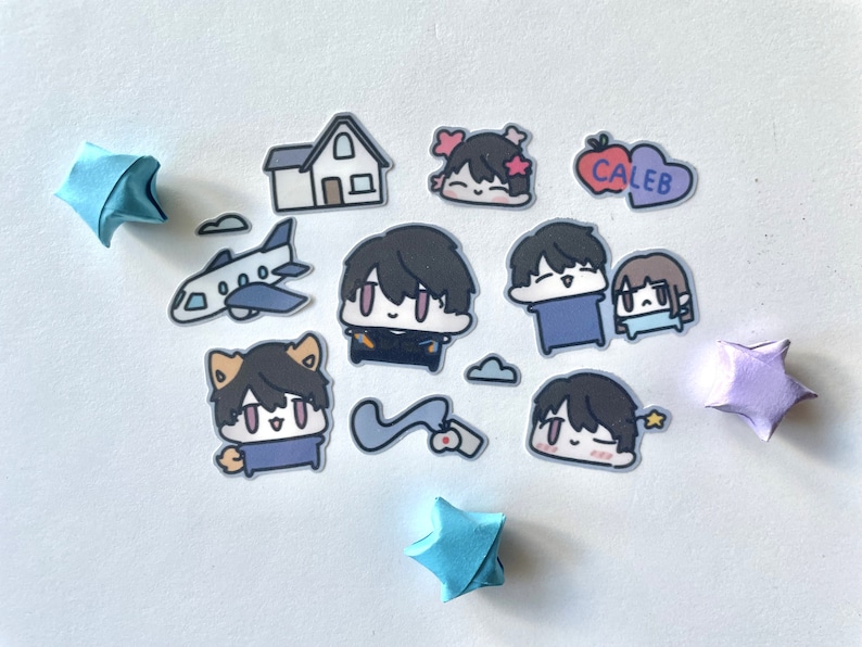 Op de afbeelding: Een set stickers met cartoonfiguren met verschillende uitdrukkingen. De stickers zijn op een witte achtergrond met origami-sterren in blauw en paars. De stickers bevatten een huis, een vliegtuig, een hart met het woord "Caleb" erop en verschillende personages met verschillende uitdrukkingen.