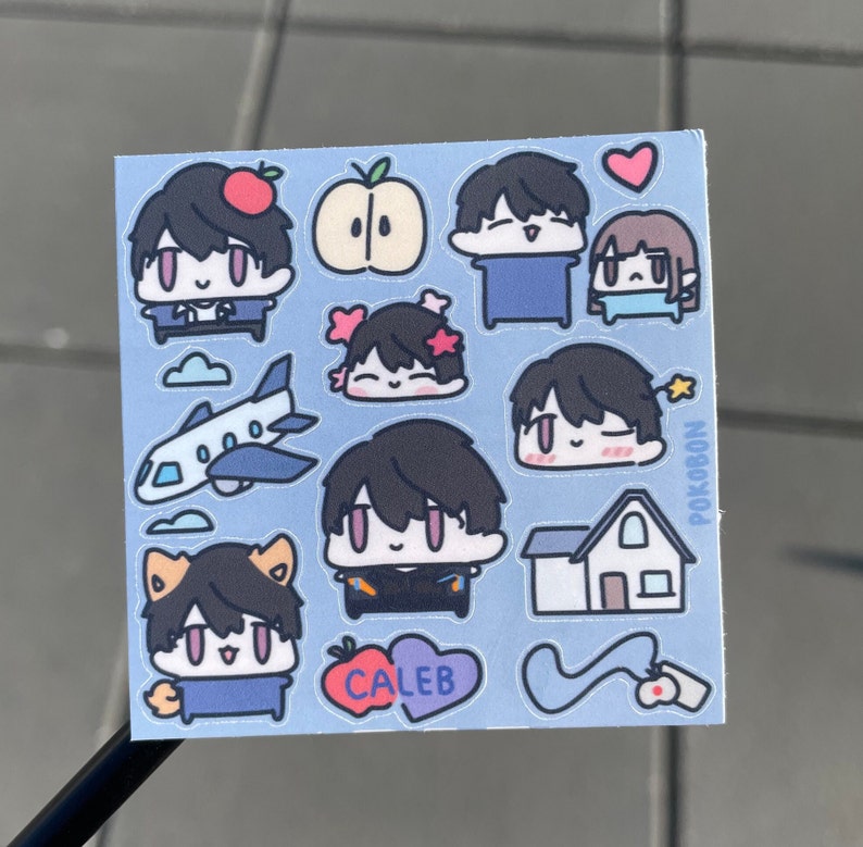 Op de afbeelding: Een vel met stickers met cartoonfiguren met verschillende gezichtsuitdrukkingen. De stickers zijn op een lichtblauwe achtergrond met witte wolken, een vliegtuig, een huis en een hart. De tekst "POKOBON" en "CALEB" is zichtbaar op de stickers.