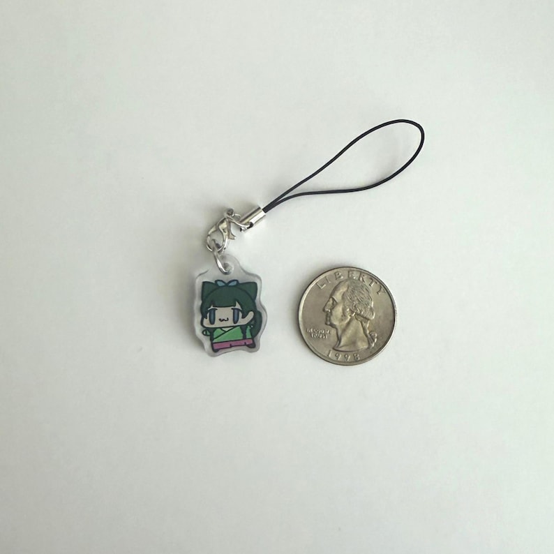 May include: A small, clear acrylic charm featuring a cartoon character with green hair and a green and pink outfit. The charm is attached to a black cord with a silver-colored clasp. A US quarter is next to the charm for size comparison.