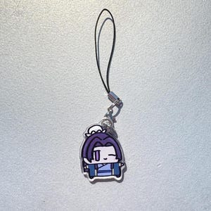 May include: A cartoon charm featuring a character with purple hair and blue clothing. The charm is attached to a black lanyard with a silver-colored clasp. The character has a white outline.