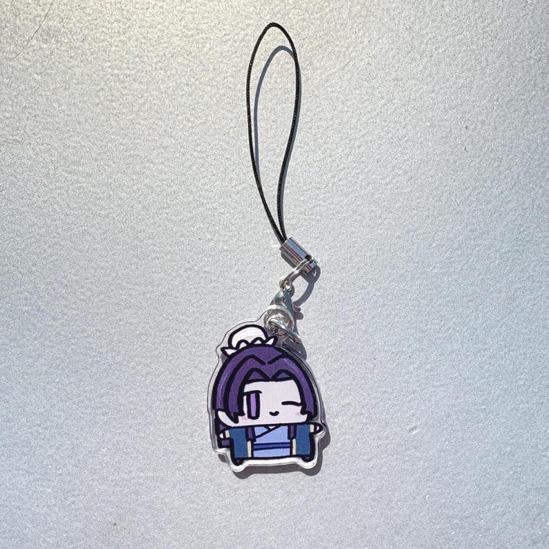 May include: A cartoon charm featuring a character with purple hair and blue clothing. The charm is attached to a black lanyard with a silver-colored clasp. The character has a white outline.