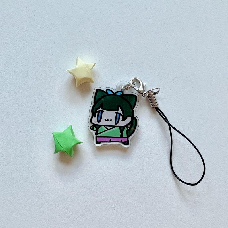 May include: A clear acrylic charm featuring a cartoon character with green hair and a cat-like design, wearing a green and purple outfit. The charm has a silver clasp and a black lanyard. Two origami stars, one yellow and one green, are also present.