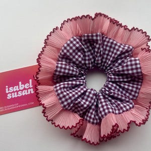 Puede incluir: Una scrunchie hecha a mano con un estampado de cuadros en el centro, rodeada de capas de tela rosa con un borde con volantes. Una tarjeta de visita con el texto "isabel susan" está al lado de la scrunchie.