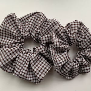 Puede incluir: Dos scrunchies de gingham marrón y blanco. Los scrunchies están hechos de tela y tienen un patrón a cuadros.