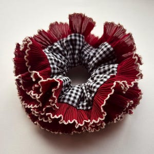 Puede incluir: Una goma para el pelo con volantes de tela plisada roja y un estampado de cuadros blancos y negros. Los bordes están ribeteados con un ribete ondulado de color crema. Este accesorio para el cabello añade un toque vintage.