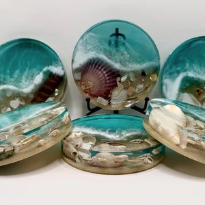 May include: Set of six round turquoise blue resin coasters with embedded seashells and sand. The coasters have a white wave design and are perfect for adding a coastal touch to your home decor.