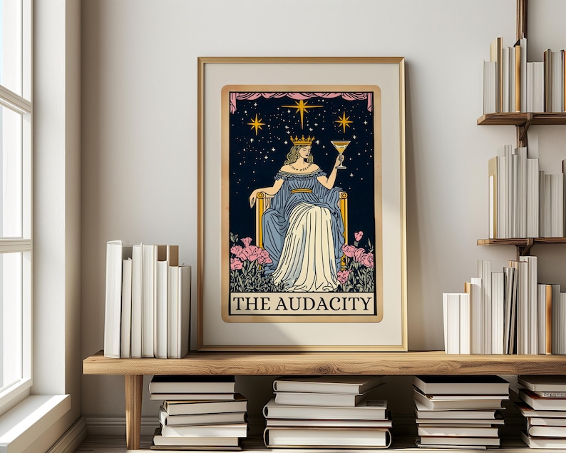 Puede incluir: Impresi&oacute;n art&iacute;stica enmarcada de una carta del tarot que representa una figura con un vestido azul sosteniendo una copa de c&oacute;ctel sobre un fondo de noche estrellada. El texto "THE AUDACITY" est&aacute; en la parte inferior. La obra de arte con marco dorado se exhibe en una estanter&iacute;a de madera con libros.
