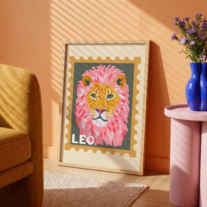 May include: A framed art print featuring a colorful illustration of a lion. The lion has a pink mane and is set against a green background within a postage stamp design. The word "LEO" is printed at the bottom. The frame is light wood.