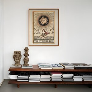Peut inclure: Une impression encadr&eacute;e d'une carte de tarot repr&eacute;sentant une femme assise en position de lotus sur une &eacute;tendue d'eau. Le soleil est au-dessus d'elle avec un visage et des rayons. La carte est intitul&eacute;e "The Overthinker". L'impression est sur une &eacute;tag&egrave;re avec des livres et des sculptures d&eacute;coratives.