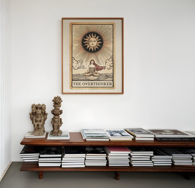 Peut inclure: Une impression encadr&eacute;e d'une carte de tarot repr&eacute;sentant une femme assise en position de lotus sur une &eacute;tendue d'eau. Le soleil est au-dessus d'elle avec un visage et des rayons. La carte est intitul&eacute;e "The Overthinker". L'impression est sur une &eacute;tag&egrave;re avec des livres et des sculptures d&eacute;coratives.